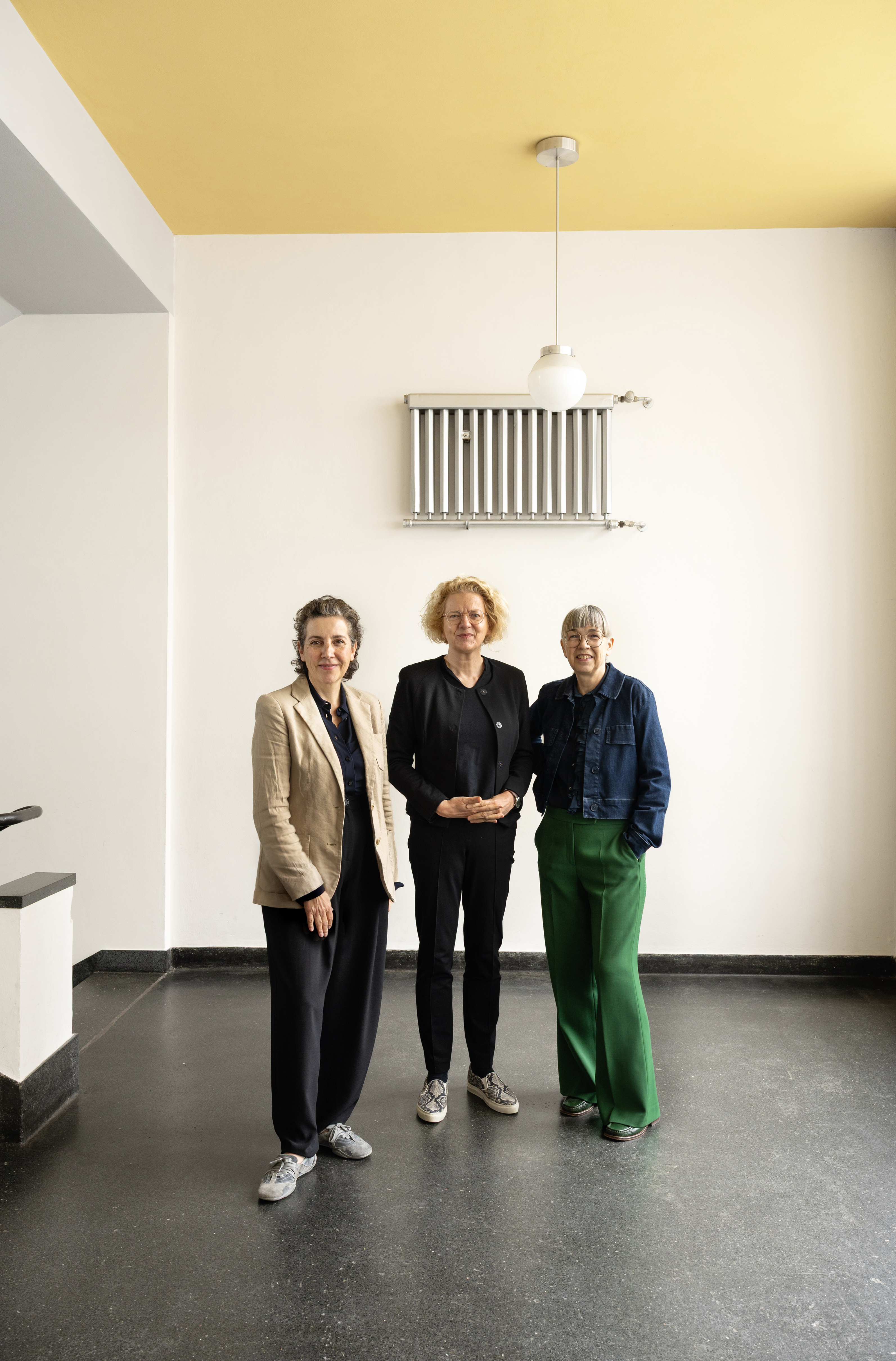 Drei Frauen stehen in einem modernen Raum mit gelbem Deckenelement und Heizkörper, alle in stilvollen Outfits.