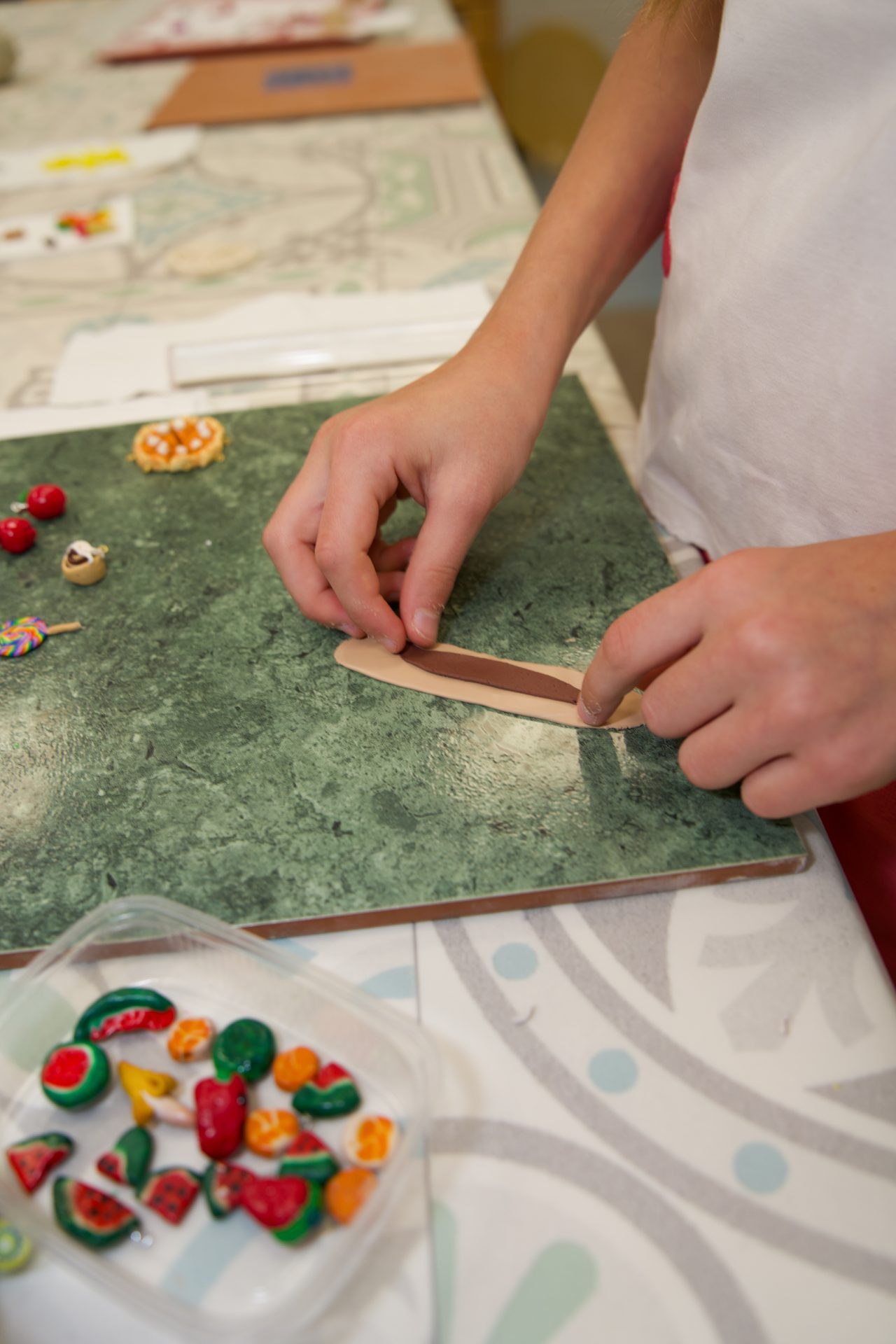 A child shapes modeling clay with their hands on a green work surface to create small food items.