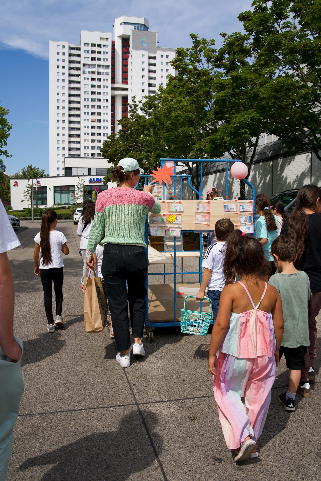 A group of children and adults walks with a cart full of goods towards a building under a blue sky.