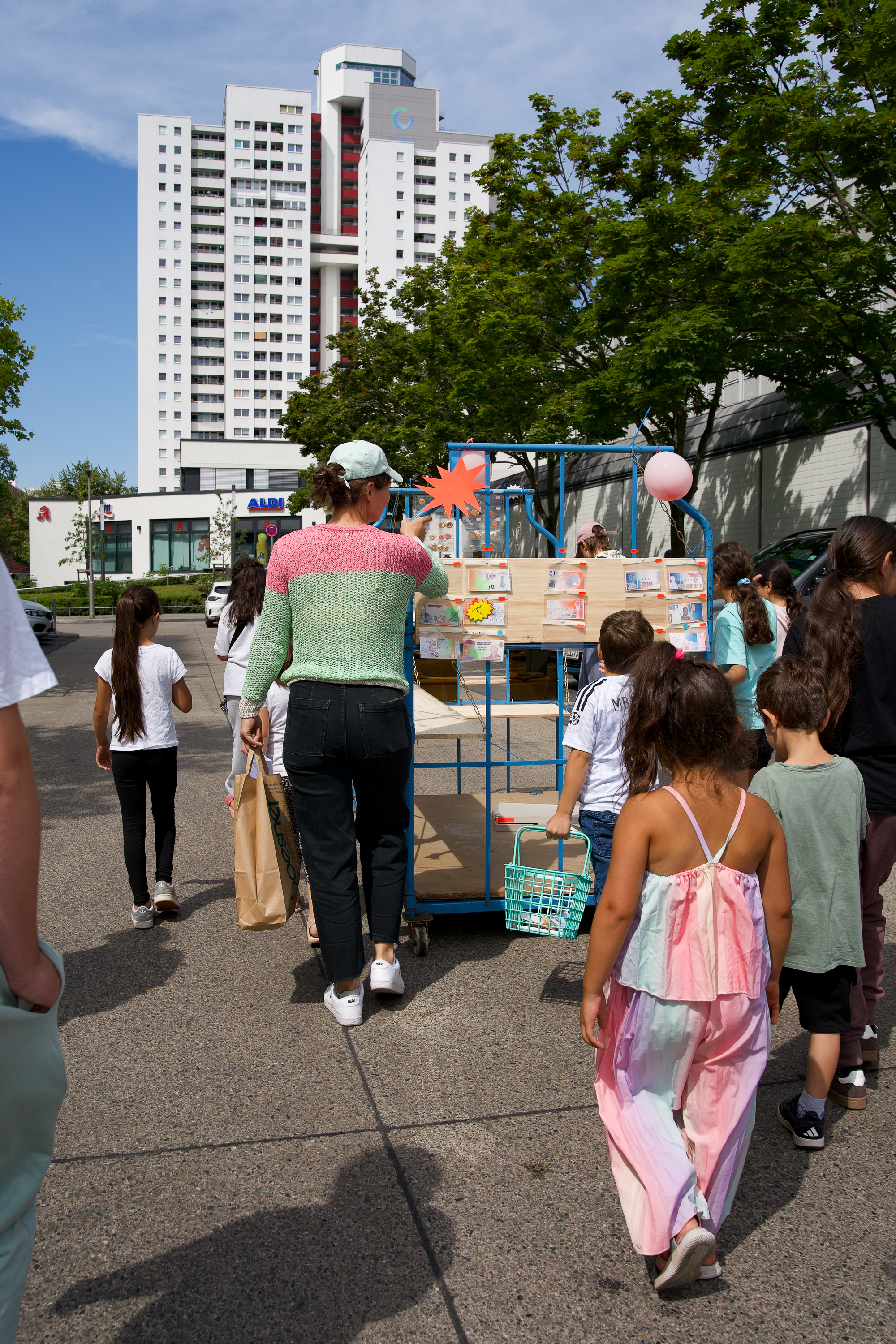 Gruppe von Kindern und Erwachsenen geht mit einem Wagen voller Waren in Richtung eines Wohngebäudes unter blauem Himmel.