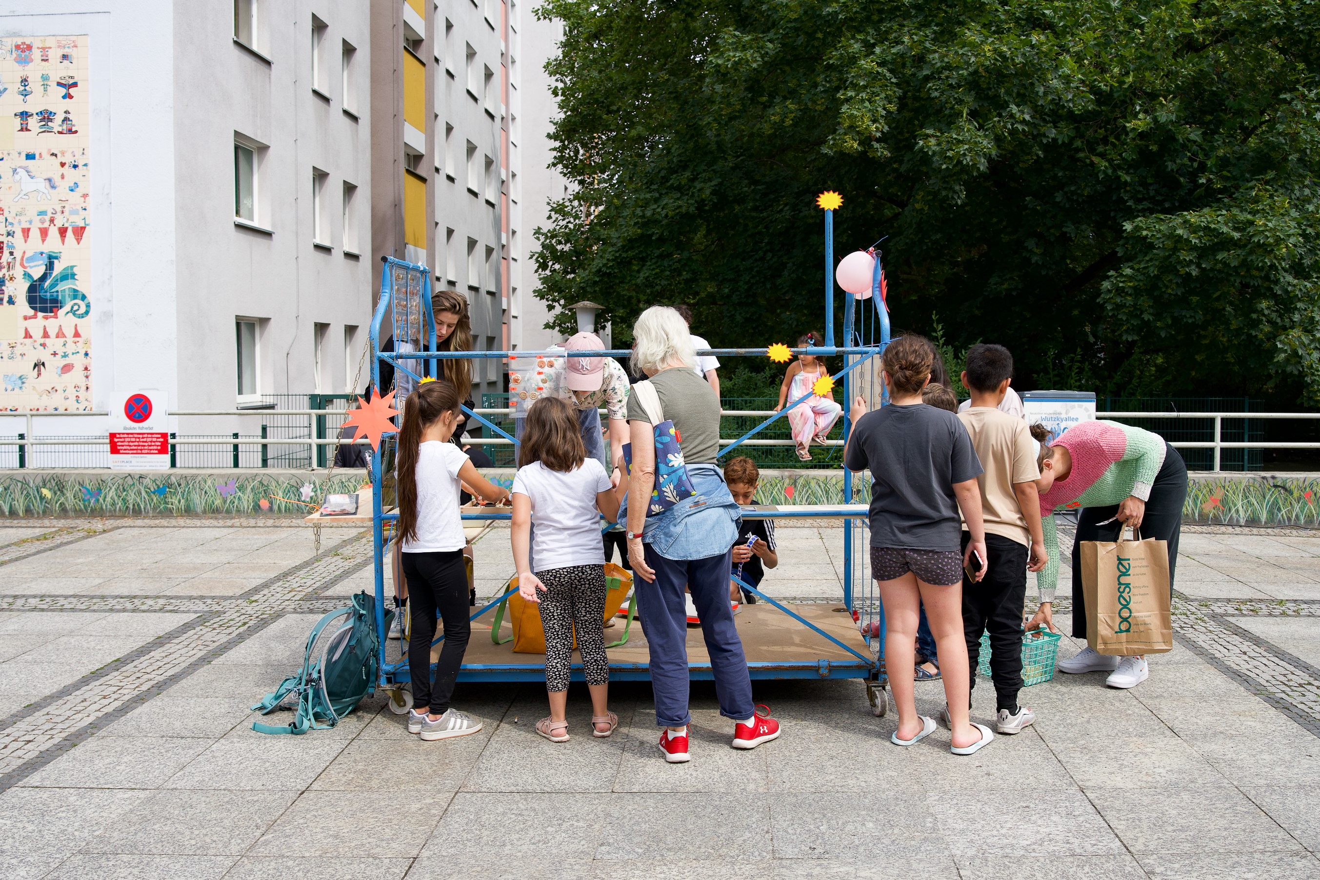 Kinder und Erwachsene spielen auf einem bunten Spielplatz mit modernen Gebäuden und viel Grün im Hintergrund.