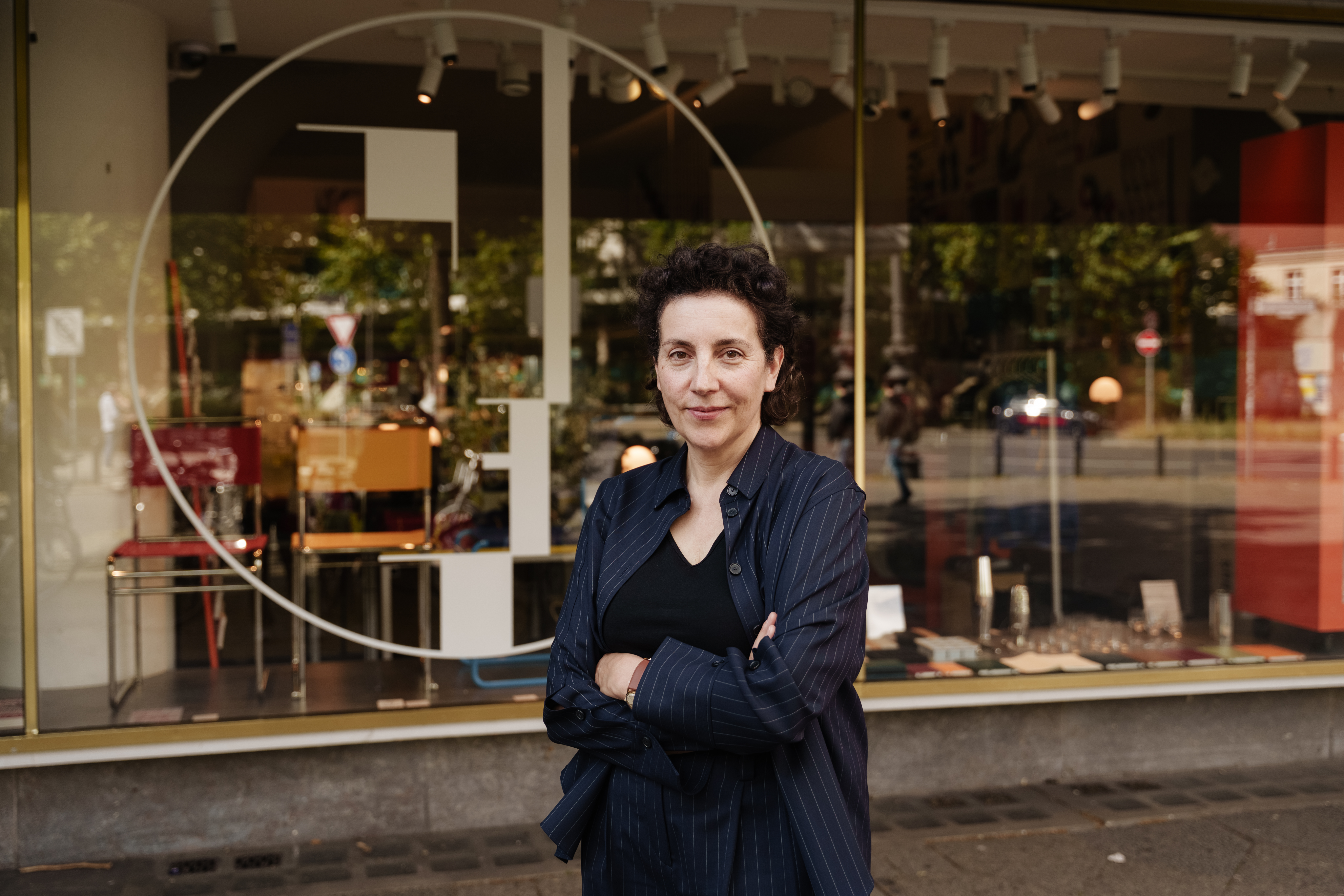 Frau in schwarzer gestreifter Bluse steht vor einem Schaufenster mit bunten Stühlen und abstraktem Design.