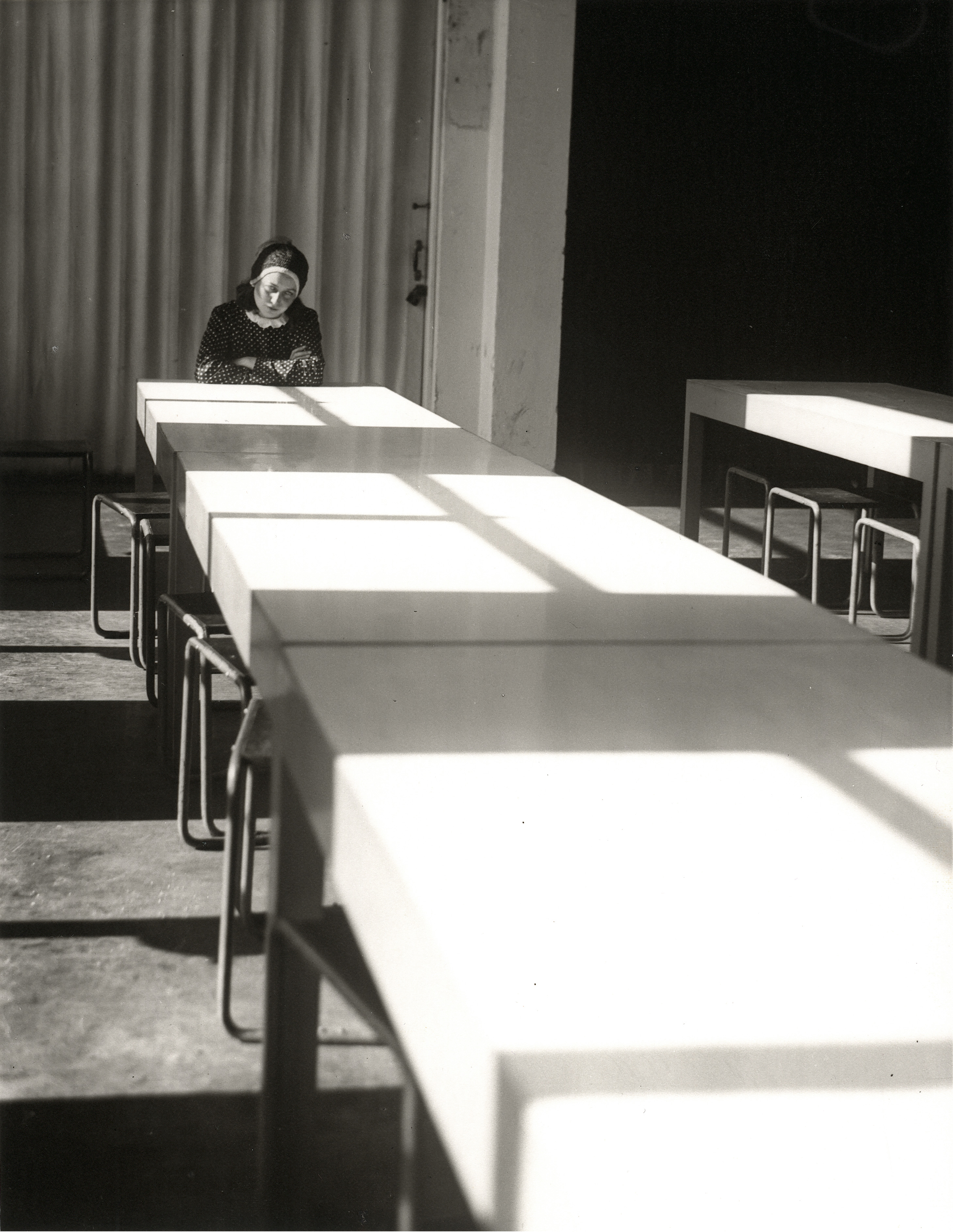 Eine Frau sitzt alleine an einem langen Tisch, umgeben von Schatten und Lichtspiel in einem minimalistischen Raum.