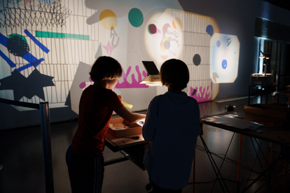 Zwei Kinder experimentieren mit einem Overheadprojektor, wobei bunte Schatten an der Wand entstehen.