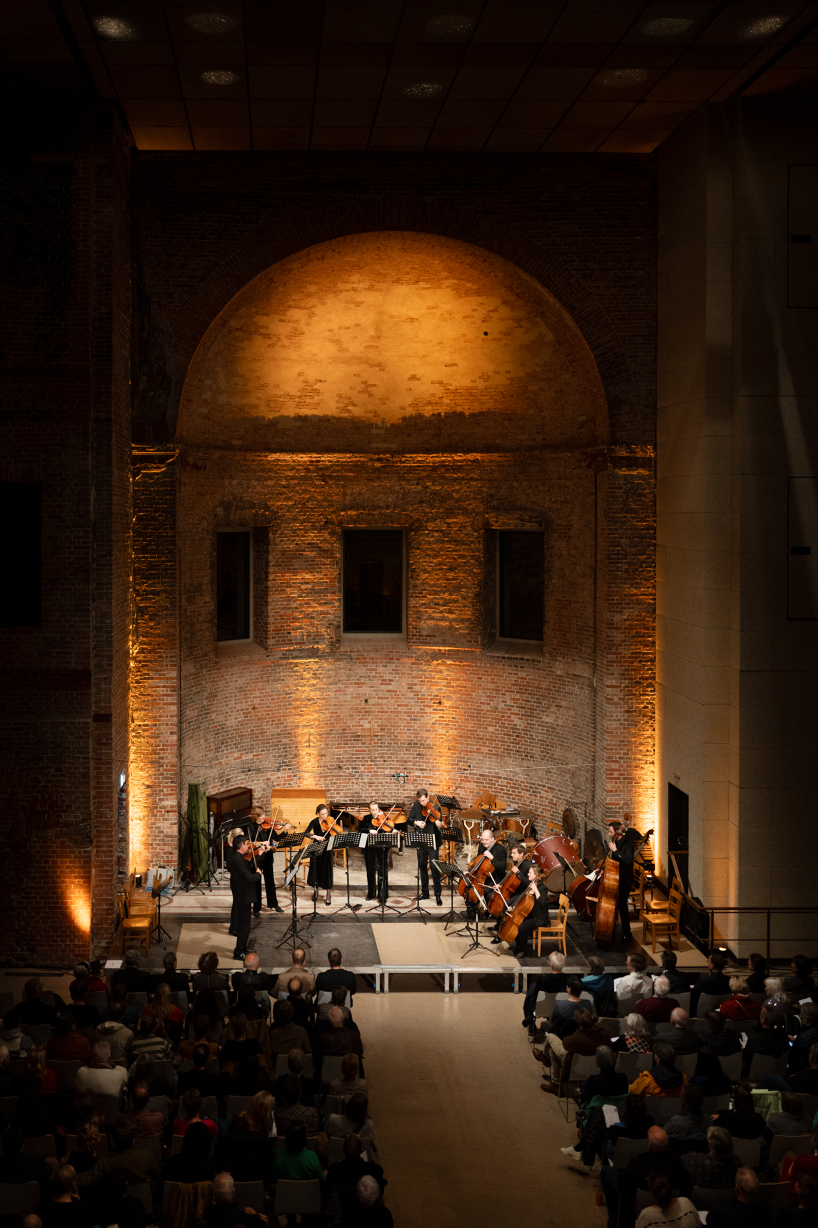Zu sehen ist der Blick in den Konzertraum einer Kirche von hinten über das Publikum hinweg bis hin zum beleuchteten Apsisbereich mit den Musiker*innen. Das Licht ist abendlich dunkel mit einer warmen Stimmung.