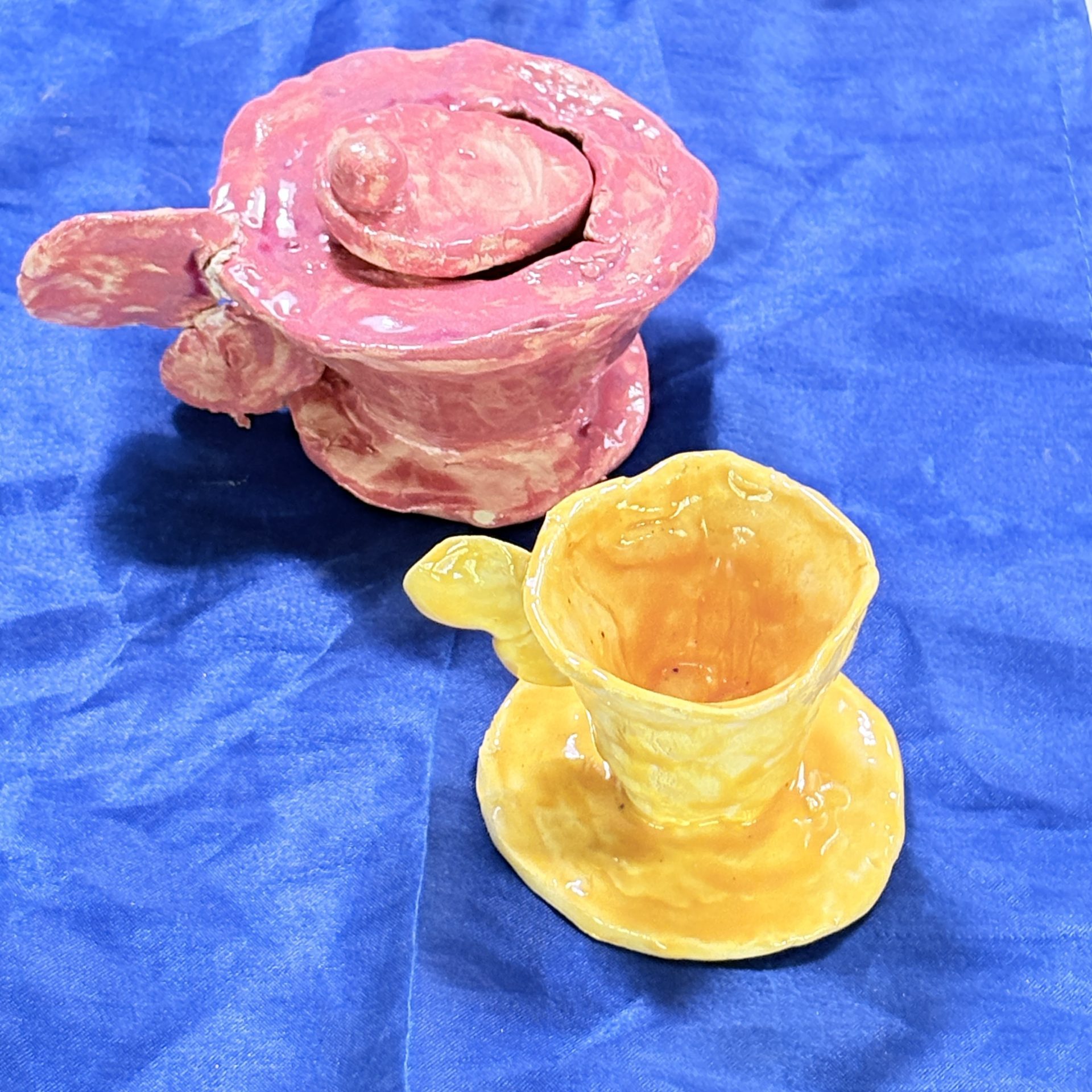 Colorful, handmade tableware: a pink teapot and an orange cup on a blue surface.
