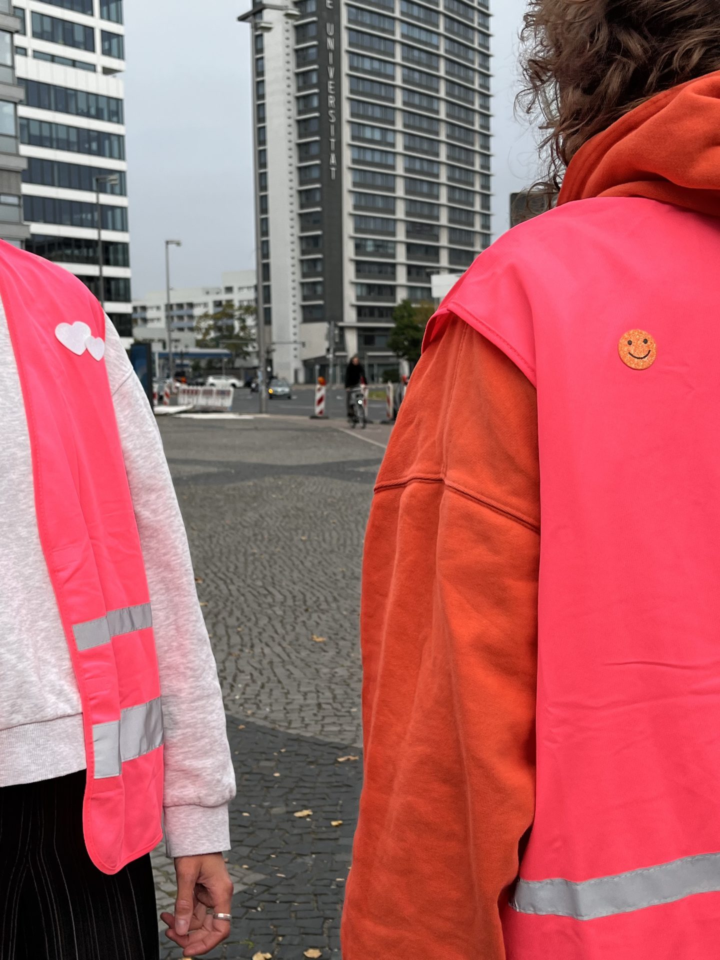 Zwei Personen mit pinkfarbenen Warnwesten über der Alltagskleidung stehen auf einem gepflasterten Platz und blicken auf zwei Hochhäuser.